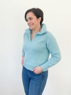 a woman standing in front of a white wall wearing a blue sweater and jeans with her hands on her hips