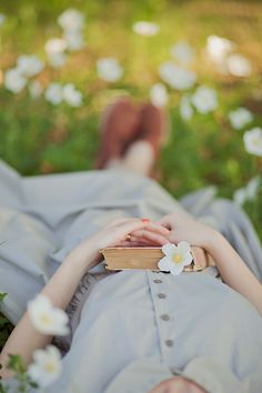 a woman laying down in the grass with a book on her lap and flowers growing out of her hair