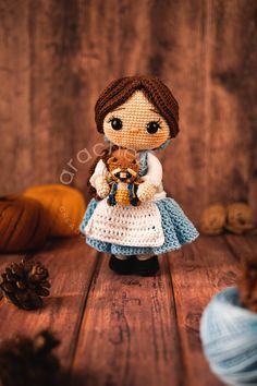 a little doll holding a teddy bear on top of a wooden table next to balls of yarn