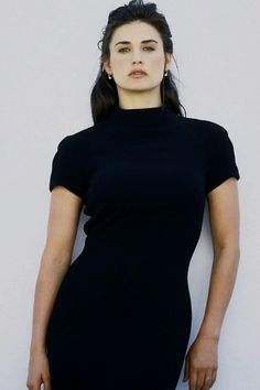 a woman in a black dress leaning against a wall with her hands on her hips