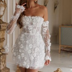 a woman in a short white dress with flowers on the sleeves and arms, standing next to a mirror