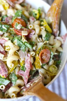 a pasta salad with ham and vegetables in a white bowl next to a wooden spoon