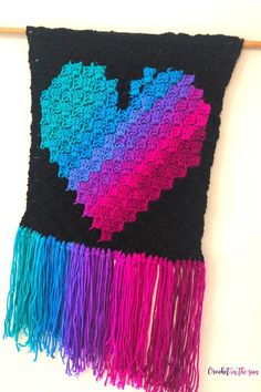 a colorful crocheted heart is hanging on a wall with fringes and yarn