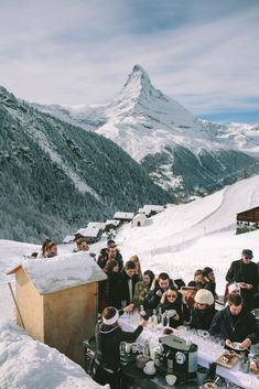 a group of people sitting at a table on top of a snow covered slope with mountains in the background