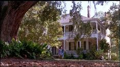 two people standing in front of a large white house with trees and leaves on the ground