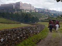 two people riding horses down a dirt path in front of a large building on top of a hill