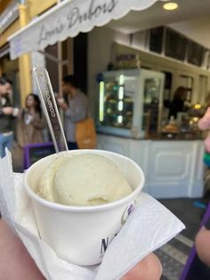 a person holding up a bowl of ice cream in front of a store with people walking by