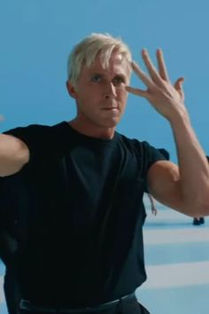 a man with blonde hair and black shirt holding his hands up in the air while standing next to a blue wall