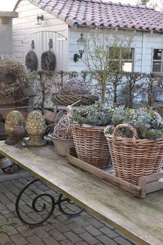 a wooden table topped with baskets filled with plants next to a white building and brick walkway