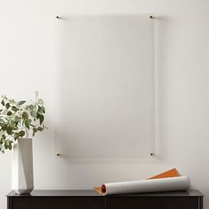 a white vase with a plant in it on top of a black dresser next to a blank canvas