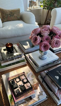 a table topped with lots of books next to a white chair and vase filled with pink roses