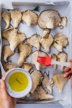 someone holding a red brush and dipping sauce into some mushrooms on a sheet of parchment paper