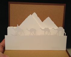 a person holding up a card with some paper cut out of it and camels in the background