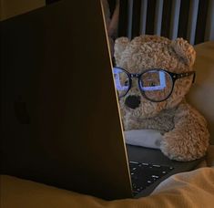 a teddy bear wearing glasses sitting in front of a laptop on a bed with sheets