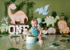 a baby sitting on the floor in front of a cake with dinosaur decorations behind it