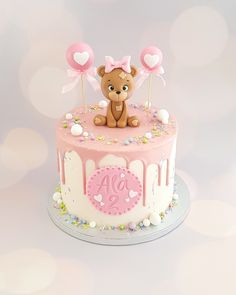 a pink and white birthday cake with a teddy bear on top that has hearts attached to it