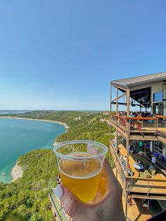 a person holding up a glass of beer on top of a building overlooking the ocean