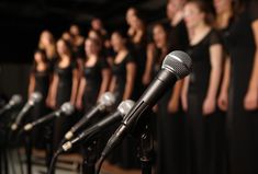 several microphones are lined up in front of a row of women singing with their hands behind them