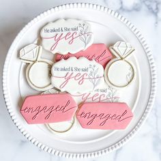 decorated cookies on a white plate with pink and white lettering that read engaged, yes asked & she said engaged