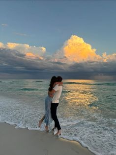 two people hugging each other on the beach near the ocean at sunset with clouds in the sky