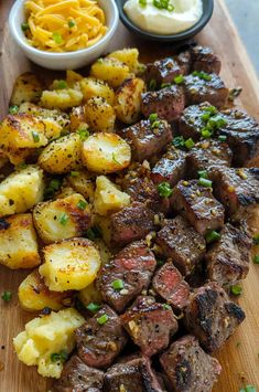 a wooden cutting board topped with steak and potatoes next to bowls of macaroni and cheese