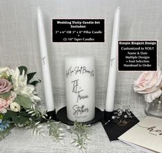 two white candles are sitting on a table with flowers and greeting cards next to it