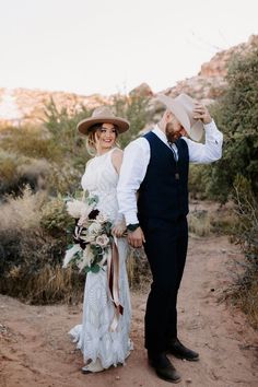 a bride and groom walking down a dirt path in the desert holding each other's hands