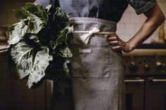 a woman wearing an apron holding a large green plant in her hands and standing next to a stove