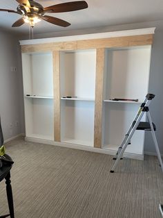 an empty room with white shelves and a ladder in the middle, next to a ceiling fan