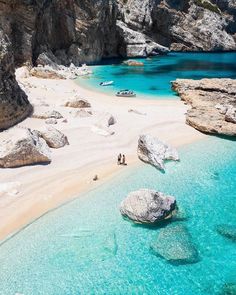 two people are walking on the beach next to some rocks and clear blue ocean water