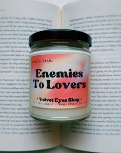 an open book is next to a jar of scented candles that reads, enemies to lovers