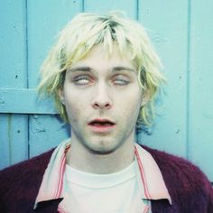 a young man with blonde hair and blue eyes wearing a pink shirt is standing in front of a blue wall