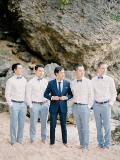 a group of men standing next to each other on top of a sandy beach under a large rock