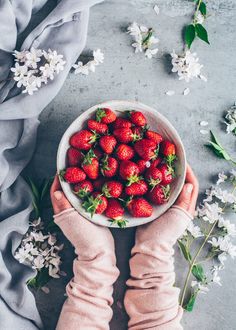 a person holding a bowl of strawberries on top of a table with white flowers