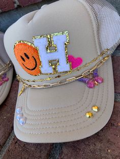 This custom hat is one of my favorite creations!! It comes with two hat chains. They are interchangeable and removable with clasps on each side. In the personalization box please state the letter patch that you would like.  If there is something on the hat that you would like changed please states that as well. For example, if you didn't want the studs on the bib or didn't want all three patches. We also have more colors in our smiley face patches. If you are interested in another color please send me a message and we can create something!  Please allow up to a week for your order to ship as this is a custom piece. Thank you for shopping Chloe Beth Designs! Customized Trucker Hats, Trendy Gold Hat As A Gift, Personalized Trucker Hat Gift, Custom Personalized Trucker Hat As Gift, Custom Adjustable Snapback Hat Gift, Gold Adjustable Trucker Hat Trendy, Custom Adjustable Trucker Hat For Gift, Trendy Personalized Adjustable Baseball Cap, Trendy Adjustable Gold Baseball Cap