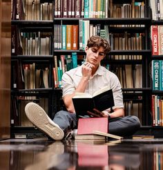 a man sitting on the floor in front of a bookshelf reading a book