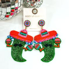 Green chili pepper beaded earrings. This pepper has arms holding colorful maracas, has a black stitched mustache, and a colorful sombrero with beaded tassels. These earrings are pictured on a white background with disco balls in the top left corner. Green Chili Peppers, Giddy Up Glamour, Plain Dress, Backyard Party, Chili Pepper, Seed Bead Earrings, Beaded Earrings, Sale Items, Seed Beads