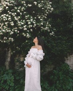 a pregnant woman in a white dress standing next to a bush with flowers on it