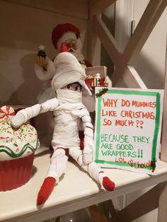 an elf is sitting next to a cupcake with a sign that says why do mummies like christmas so much? because they are good wrappers