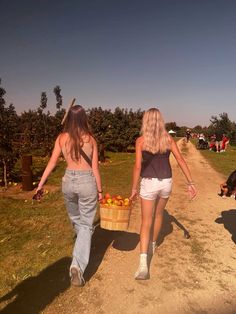 two women walking down a dirt road carrying baskets of apples in each hand and an orange on the other