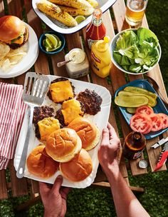 a person holding a plate with hamburgers and other food on it next to corn