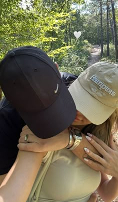 two people are hugging each other in the woods with trees and leaves behind them, one is wearing a hat