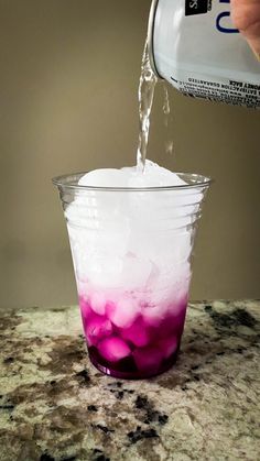 a person pours water into a cup filled with ice and purple liquid on a marble countertop