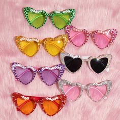 six pairs of sunglasses with different colors and shapes on pink furnishing, including one in the shape of a heart