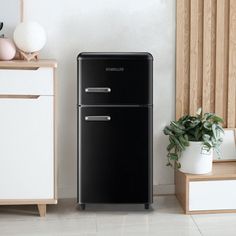 a black refrigerator sitting next to a plant on top of a wooden shelf in front of a white wall