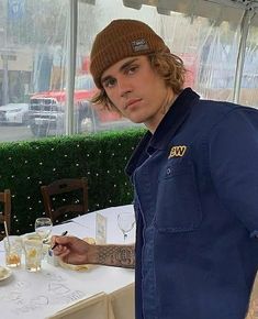 a man with long hair wearing a blue shirt and hat standing in front of a table