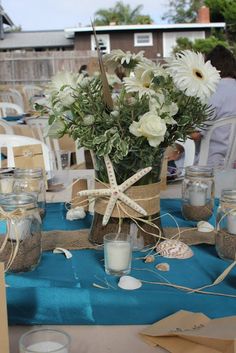 a table topped with mason jars filled with flowers and seashells on top of a blue cloth covered table