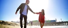 a man and woman holding hands while walking on the sand at the beach in front of a pier