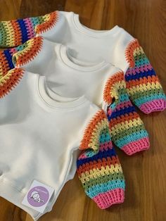 three baby onesuits with colorful crocheted sleeves on top of a wooden floor