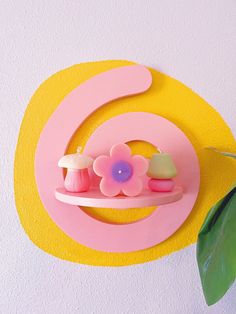 a pink and yellow wall hanging on the side of a white wall next to a green plant